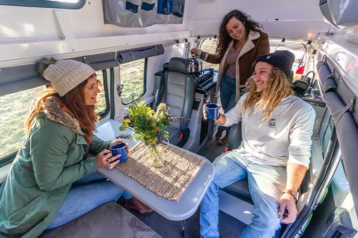 Three friends laughing over coffee while sitting at the table set up inside their JUCY Campervan