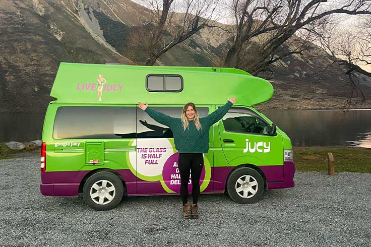 Girl standing in front of a JUCY Condo parked beside a lake and mountain view